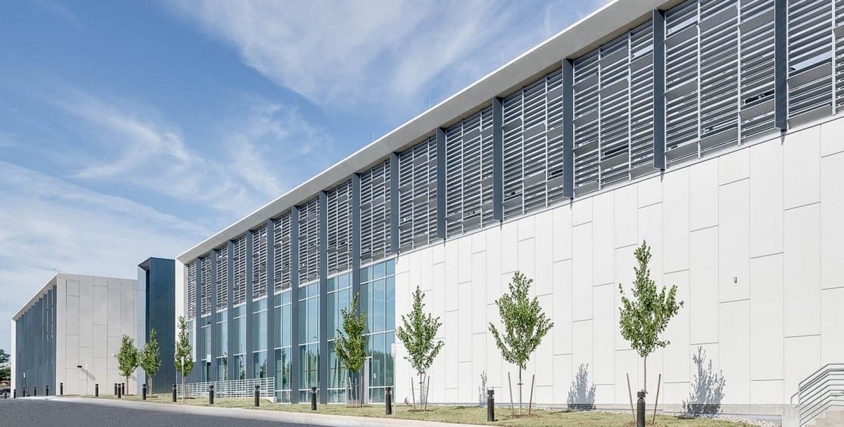 Exterior view of a modern data center building with glass panels, metal louvered screens, and young trees along the sidewalk.