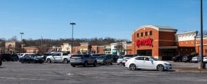 Shopping center parking lot with multiple vehicles parked near retail stores under a clear sky.